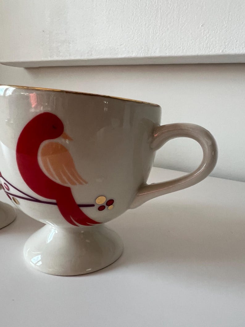 May include: A white ceramic mug with a gold rim and a red and orange bird design. The mug has a pedestal base.