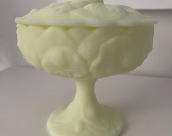 Op de afbeelding: Een lichtgele, afgedekte bonbonschaal met een voet. De schaal en het deksel zijn versierd met een bloemmotief. Het deksel heeft een kleine, decoratieve knop. De schaal is van glas.