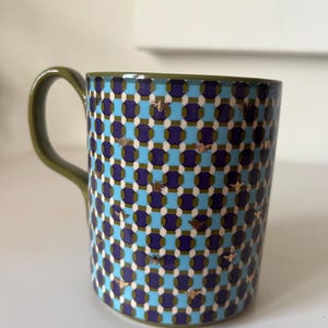 Puede incluir: Una taza de cer&aacute;mica con asa y borde verdes. La taza presenta un patr&oacute;n repetitivo de cuadrados azules, morados y blancos, con detalles de abejas doradas. La taza est&aacute; dise&ntilde;ada para bebidas fr&iacute;as o calientes.