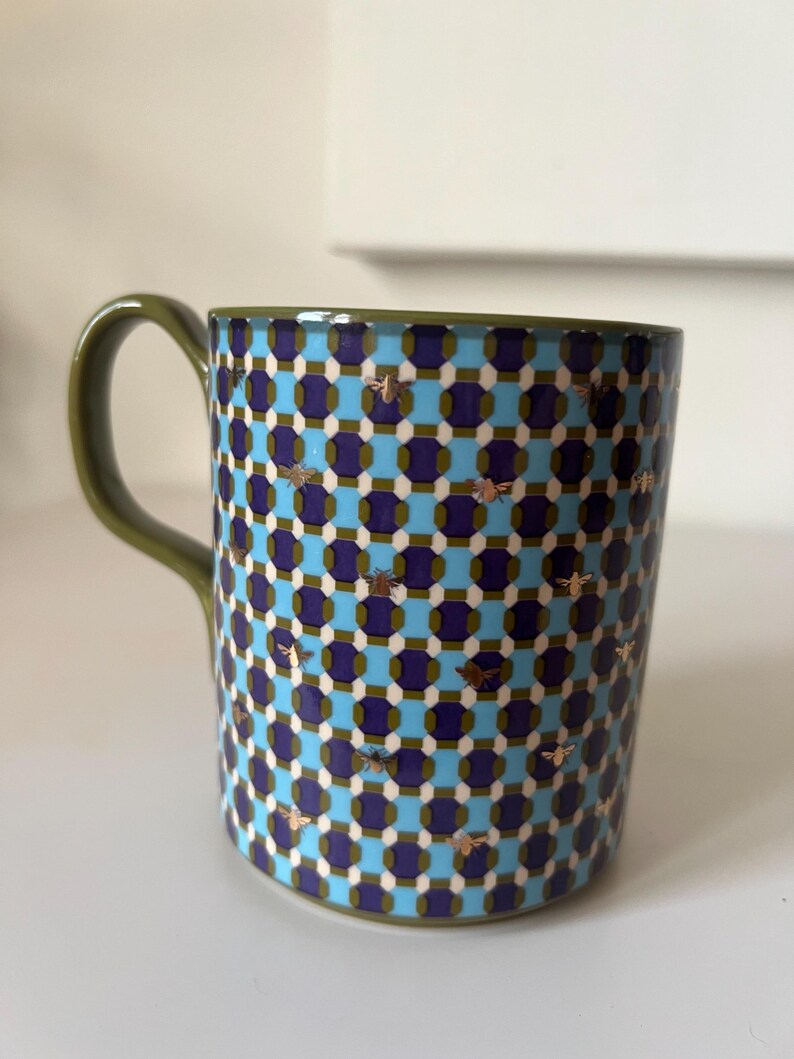 Puede incluir: Una taza de cer&aacute;mica con asa y borde verdes. La taza presenta un patr&oacute;n repetitivo de cuadrados azules, morados y blancos, con detalles de abejas doradas. La taza est&aacute; dise&ntilde;ada para bebidas fr&iacute;as o calientes.