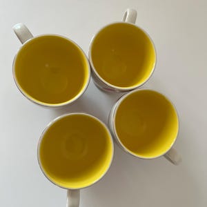 May include: Four white ceramic mugs with yellow interiors. The mugs have a simple design and are perfect for everyday use.