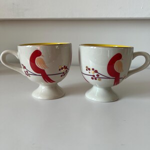 May include: Two white ceramic mugs with yellow rims and red bird designs. The birds are perched on branches with red berries and gold accents.