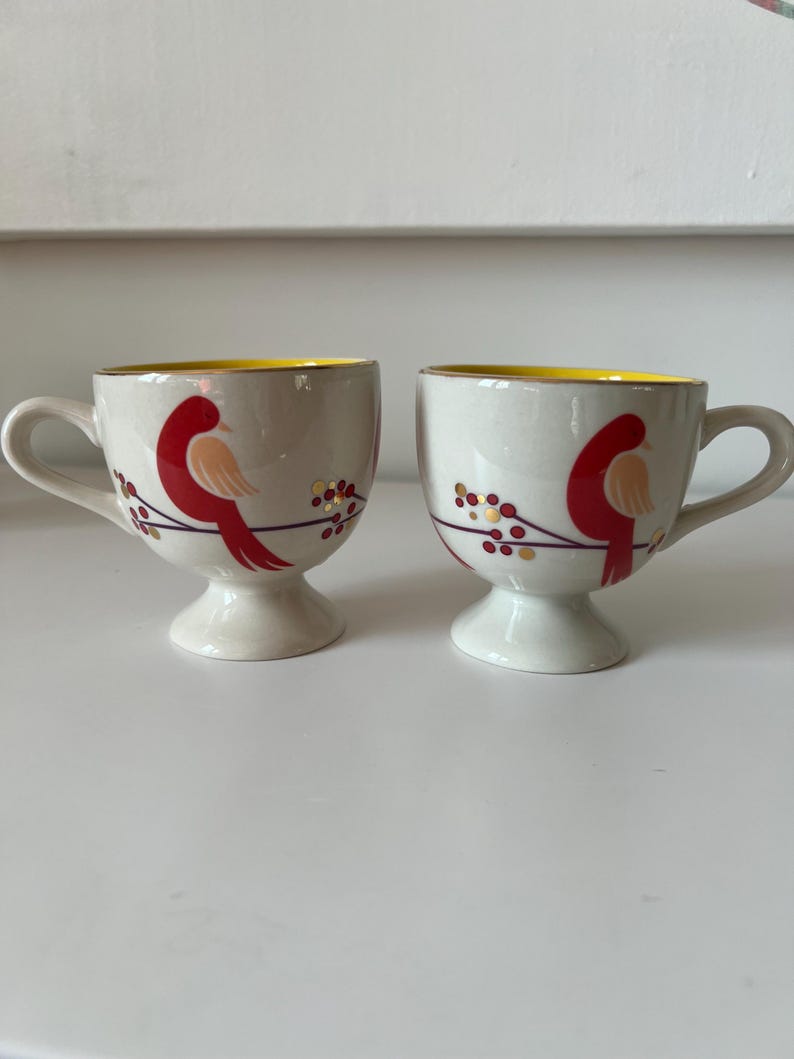 May include: Two white ceramic mugs with yellow rims and red bird designs. The birds are perched on branches with red berries and gold accents.