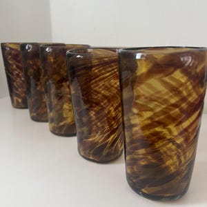 May include: Six brown and amber-colored drinking glasses. The glasses have a cylindrical shape with a textured, swirled pattern. The glasses are arranged in a row, showcasing their unique design and color variations. Ideal for serving beverages.