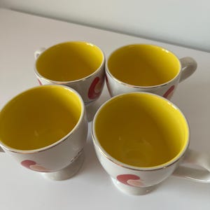 May include: Set of four white ceramic mugs with yellow interiors and red, pink, and gold abstract designs. The mugs have a rounded shape and a small foot.