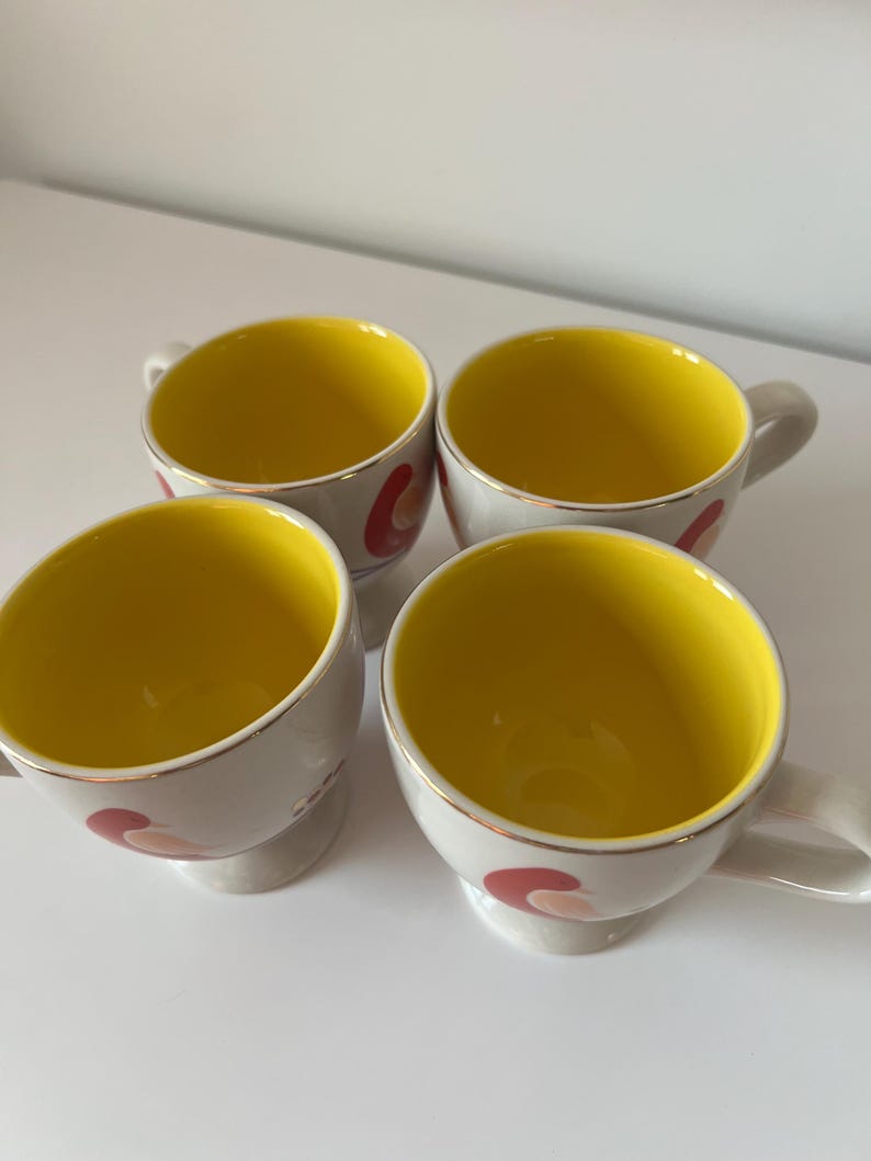 May include: Set of four white ceramic mugs with yellow interiors and red, pink, and gold abstract designs. The mugs have a rounded shape and a small foot.