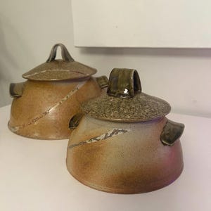 May include: Two handmade ceramic lidded jars. The jars are a mix of tan and brown with a speckled lid and a curved handle. Each jar has a diagonal white line design and small side handles.