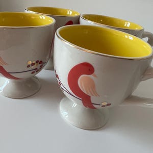 May include: Set of four ceramic mugs with a white base and yellow interior. Each mug features a red bird with gold accents and a gold rim. The mugs have a pedestal base.
