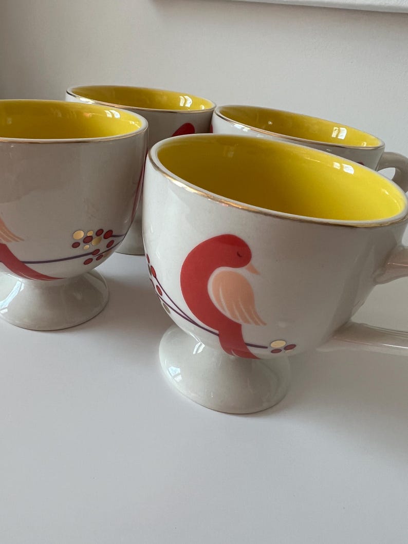 May include: Set of four ceramic mugs with a white base and yellow interior. Each mug features a red bird with gold accents and a gold rim. The mugs have a pedestal base.