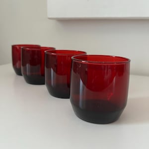 May include: Four red drinking glasses with a dark base are arranged in a row. The glasses have a rounded shape and a smooth, glossy finish. The background is a plain white surface.