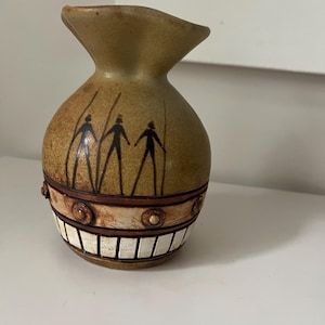 May include: A tan ceramic vase with a flared top and a rounded body. The vase features a band of decorative elements, including small circles and rectangular shapes. Three stick figures are painted on the front of the vase.