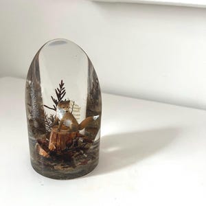 May include: A clear resin paperweight featuring a miniature woodland scene. A small fox sits atop a wooden stump, surrounded by miniature trees and foliage. The resin is clear, allowing a full view of the detailed diorama.