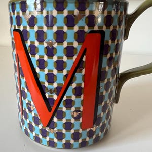 Puede incluir: Una taza de cer&aacute;mica con un patr&oacute;n geom&eacute;trico en azul, morado y blanco, con una gran letra "M" naranja con contorno negro. La taza tiene un asa verde y est&aacute; decorada con peque&ntilde;os motivos de abejas doradas.