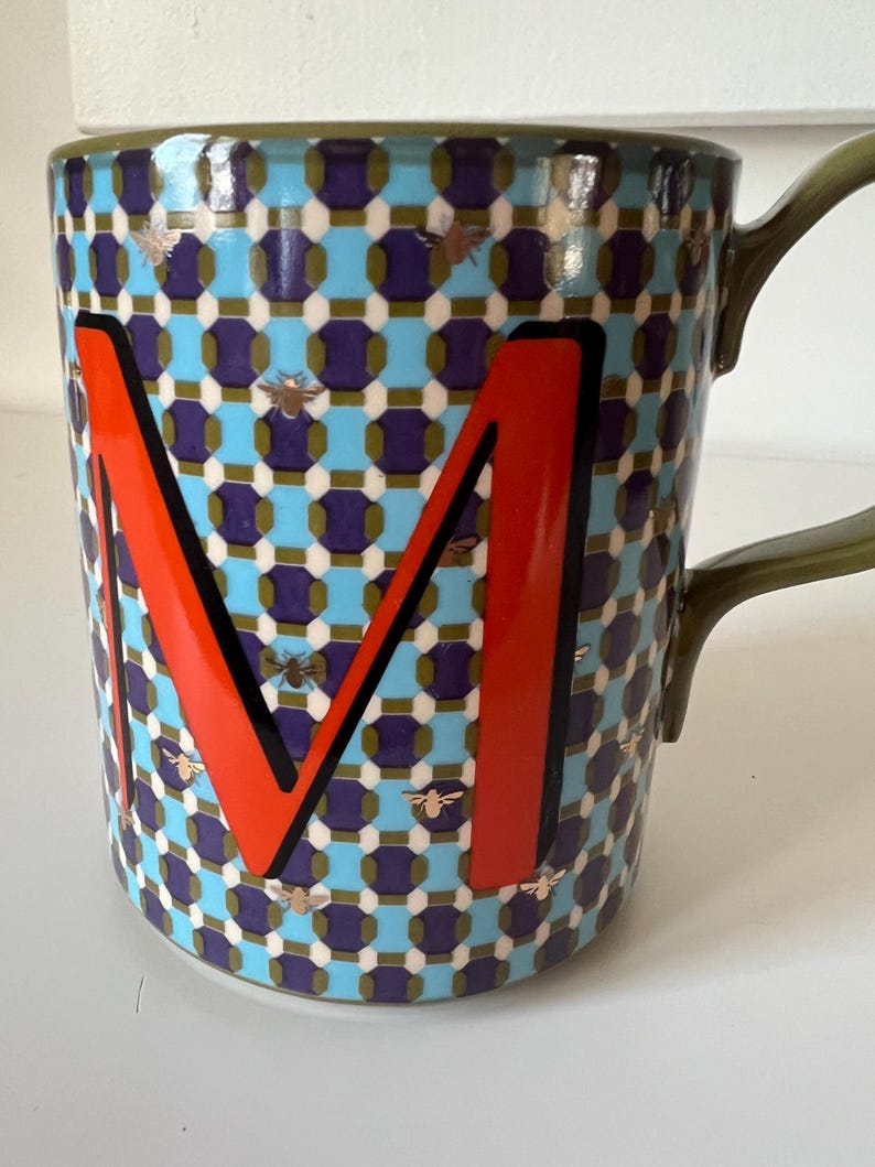 Puede incluir: Una taza de cer&aacute;mica con un patr&oacute;n geom&eacute;trico en azul, morado y blanco, con una gran letra "M" naranja con contorno negro. La taza tiene un asa verde y est&aacute; decorada con peque&ntilde;os motivos de abejas doradas.