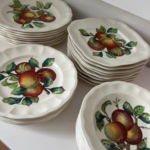 May include: A collection of white ceramic plates with a scalloped edge. Each plate features a detailed illustration of ripe peaches with green leaves. The plates are stacked in multiple piles, showcasing a classic fruit design.