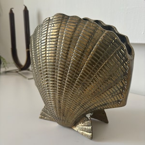 May include: A gold-colored brass seashell planter with a detailed, textured design. The planter is shaped like a large seashell with a wide opening and a flat base.