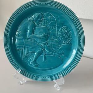 May include: A turquoise ceramic plate with a raised design of a person working on a pottery wheel. The plate has a decorative border of small dots.