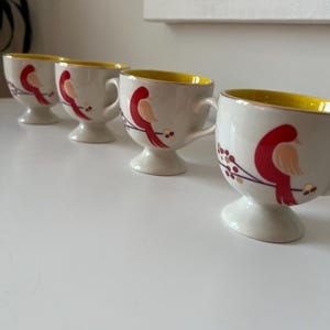 May include: Set of four white ceramic mugs with yellow rims and red bird illustrations. Each mug has a pedestal base.