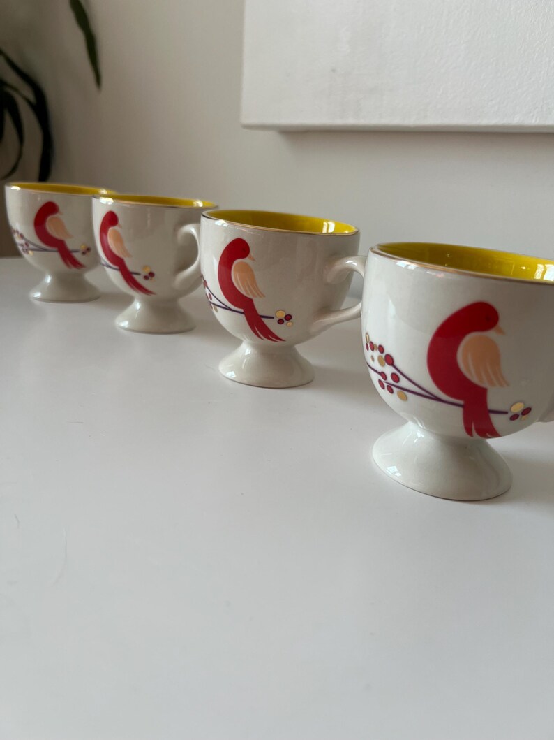 May include: Set of four white ceramic mugs with yellow rims and red bird illustrations. Each mug has a pedestal base.