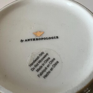 Puede incluir: Cuenco de cer&aacute;mica blanquecina con borde verde. El cuenco tiene el texto "by ANTHROPOLOGIE" y un peque&ntilde;o logotipo de abeja dorada. Una pegatina en el cuenco dice "Handwash Only Do Not Microwave Made in China Fabriqu&eacute; en Chine Hecho en China."