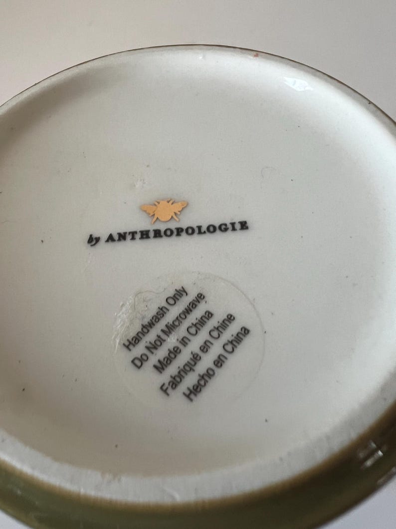 Puede incluir: Cuenco de cer&aacute;mica blanquecina con borde verde. El cuenco tiene el texto "by ANTHROPOLOGIE" y un peque&ntilde;o logotipo de abeja dorada. Una pegatina en el cuenco dice "Handwash Only Do Not Microwave Made in China Fabriqu&eacute; en Chine Hecho en China."