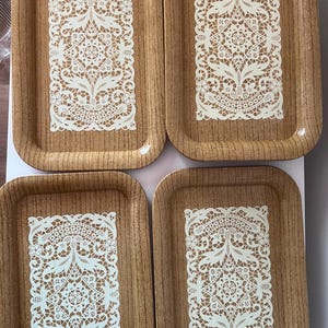 May include: Set of four wooden trays with a white lace design. The trays are rectangular and have a brown wood grain finish.