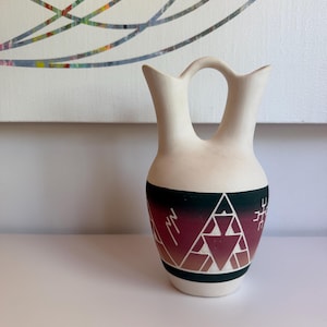 May include: A cream-colored, double-spouted vase with a handle. The vase features a geometric design in black, red, and white. The design includes triangles and linear patterns. A black band encircles the base.
