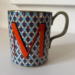 Puede incluir: Una taza de cer&aacute;mica con un patr&oacute;n geom&eacute;trico en azul, morado y blanco, con una letra "M" naranja. La taza tiene un asa y un borde verdes, y peque&ntilde;as ilustraciones de abejas. Un art&iacute;culo elegante y decorativo.