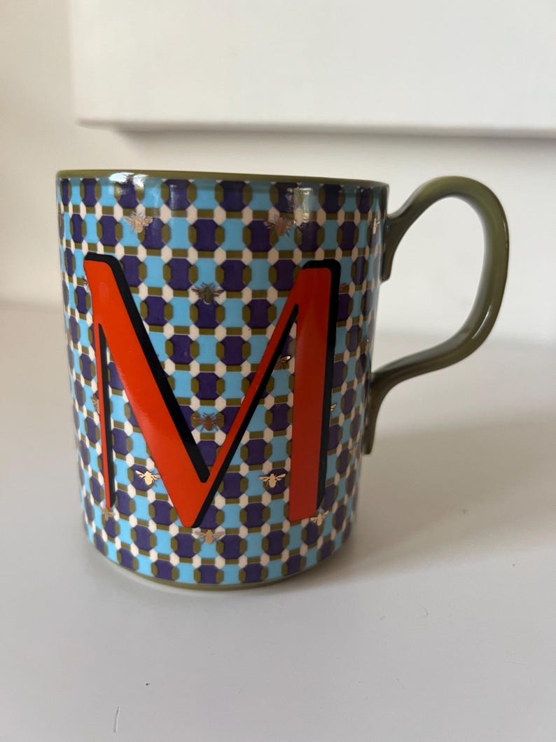 Puede incluir: Una taza de cer&aacute;mica con un patr&oacute;n geom&eacute;trico en azul, morado y blanco, con una letra "M" naranja. La taza tiene un asa y un borde verdes, y peque&ntilde;as ilustraciones de abejas. Un art&iacute;culo elegante y decorativo.