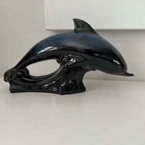May include: A glossy black ceramic dolphin sculpture with a dark blue gradient on its back and fin. The dolphin is depicted leaping over a wave, creating an open space within its form. The sculpture is a decorative item.