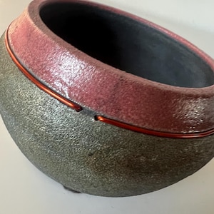 May include: A handcrafted ceramic bowl with a textured, speckled gray exterior and a reddish-pink rim. A thin, red wire encircles the bowl. The interior is dark. This decorative bowl is suitable for home decor.