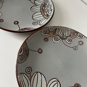 May include: Two gray ceramic plates with brown rims and floral designs. The plates feature a hand-painted design with white, teal, yellow, and brown accents. The floral pattern is a stylized, repeating motif. The plates are on a white surface.