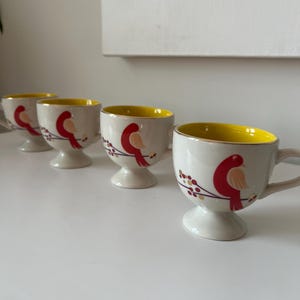 May include: Set of four white ceramic mugs with yellow rims and red bird designs. Each mug has a small foot.