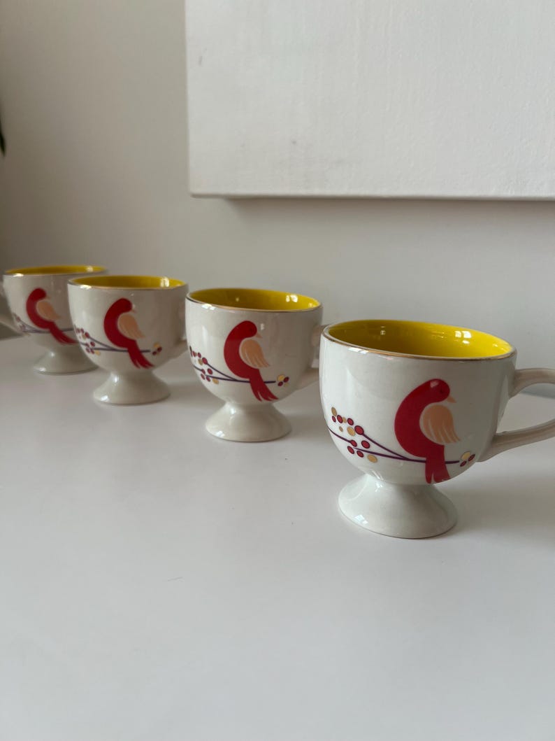 May include: Set of four white ceramic mugs with yellow rims and red bird designs. Each mug has a small foot.
