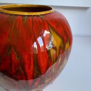 May include: A round ceramic vase with a glossy finish featuring a vibrant drip glaze pattern in shades of bright red, deep orange, and golden yellow. The vase has a wide, open mouth with a smooth, rounded rim and a textured, bulbous body.