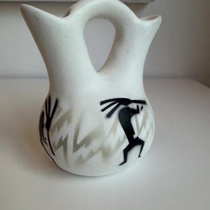 May include: A white ceramic vessel with a double-spout design and a central handle. The exterior features black painted figures of flute players and gray zigzag patterns encircling the lower body of the rounded, matte-finished container.