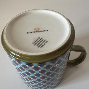 Puede incluir: Una taza de cer&aacute;mica con un patr&oacute;n geom&eacute;trico en azul, blanco y marino. El borde y el asa son de color verde oliva. La base es blanca con el texto "by ANTHROPOLOGIE" e instrucciones de cuidado. Hecho en China.