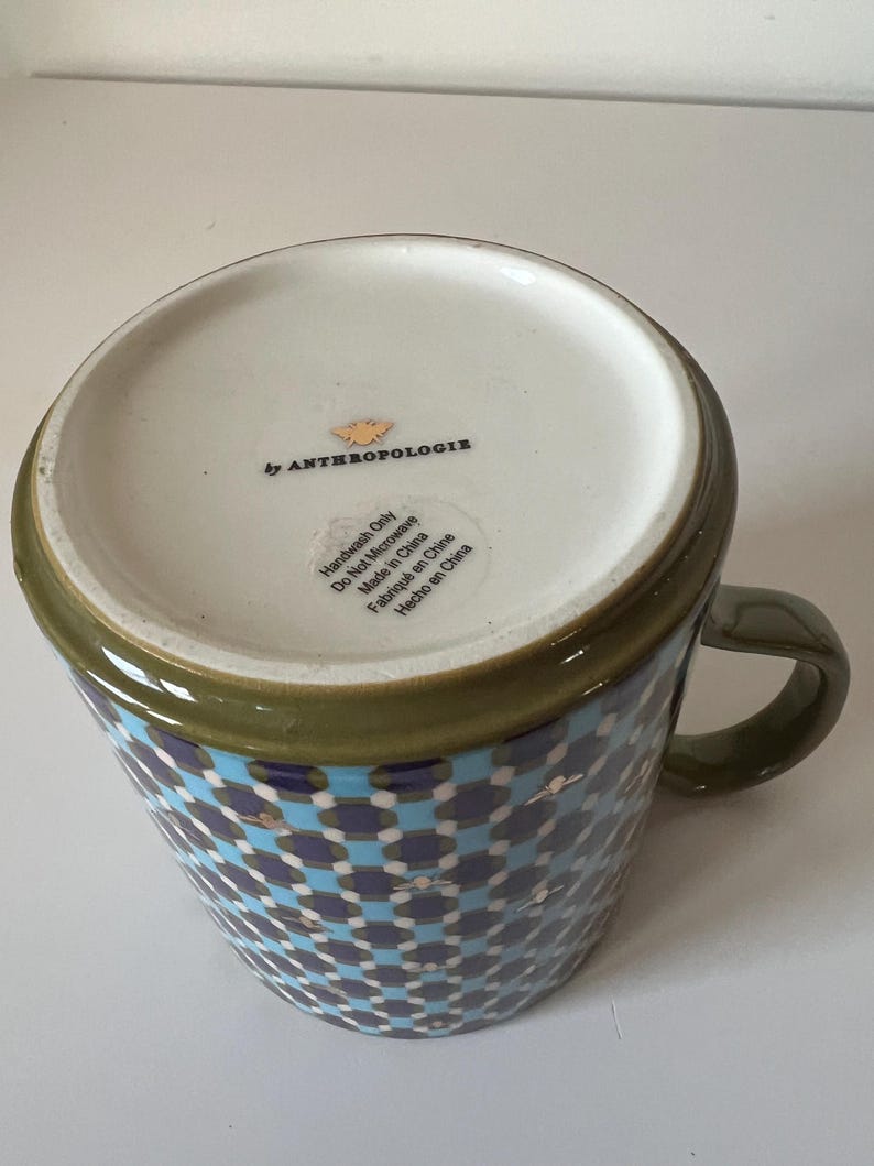 Puede incluir: Una taza de cer&aacute;mica con un patr&oacute;n geom&eacute;trico en azul, blanco y marino. El borde y el asa son de color verde oliva. La base es blanca con el texto "by ANTHROPOLOGIE" e instrucciones de cuidado. Hecho en China.
