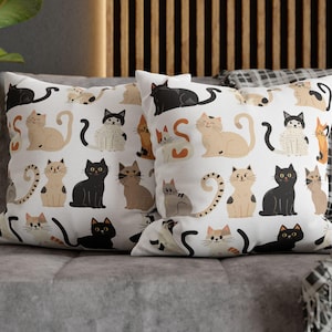 May include: Two white throw pillows with a repeating pattern of various cat illustrations in black, tan, and gray. The cats are in different poses and have unique markings. The pillows are on a gray couch.