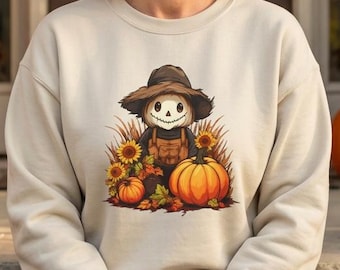 May include: A beige crewneck sweatshirt featuring a graphic illustration of a smiling scarecrow wearing a brown hat, surrounded by sunflowers and pumpkins. The design is centered on the chest of the long-sleeved garment, which is worn with dark blue denim jeans.