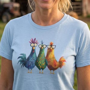 May include: A light blue short-sleeved t-shirt featuring a graphic print of three colorful chickens standing side by side. The chicken on the left is blue with a pink crest, the middle one is green, and the right one is orange wearing black glasses.