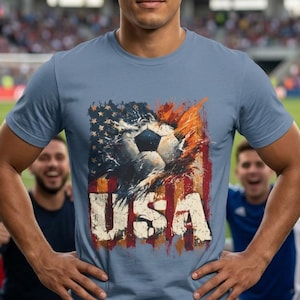 May include: A light blue t-shirt featuring a soccer ball design integrated with the American flag and the word "USA" in a distressed font. The shirt is worn by a person in a stadium setting, surrounded by cheering fans.