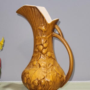 May include: A brown ceramic pitcher with a white rim and a handle. The pitcher is decorated with a grapevine design.