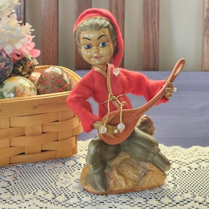 May include: A vintage figurine of a seated musician in a red hooded outfit, playing a brown lute. The figurine has a painted face with blue eyes and is seated on a textured base. The musician's outfit is trimmed with gold accents and clear beads.
