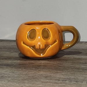 May include: Orange ceramic mug shaped like a jack-o'-lantern with a smiling face. The mug has a rounded body, a handle, and a glossy finish. Perfect for Halloween or fall-themed decor.