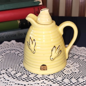 May include: A yellow ceramic pitcher with a lid and handle. The pitcher has a beehive design with three decorative bird or leaf-like accents. The pitcher is sitting on a white lace tablecloth.