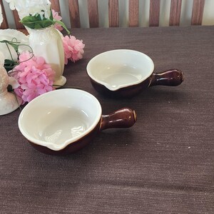 May include: Two brown and white ceramic soup bowls with handles. The bowls have a rounded shape with a pouring spout and a glossy finish. The handles are a darker shade of brown. The bowls are on a brown surface.