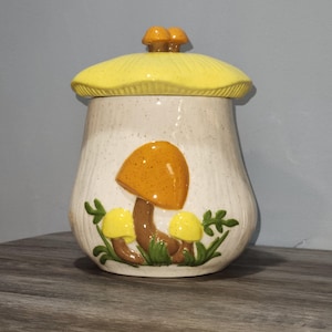 May include: A ceramic mushroom-shaped cookie jar with a yellow lid and a cluster of small mushrooms on top. The jar is off-white with brown speckles and features a large orange mushroom and two smaller yellow mushrooms with green leaves.