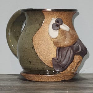 May include: Handmade ceramic mug with a whimsical bird design. The mug features a brown, tan, and green color scheme with a sculpted bird face and body. The handle is a dark green color.
