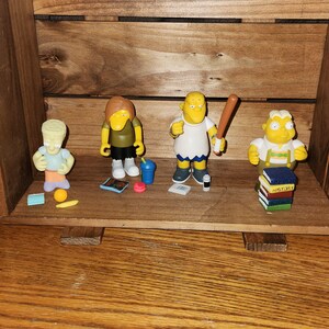 May include: Four Simpsons character figurines displayed inside a wooden crate. The figurines include Bart, Lisa, Homer holding a baseball bat, and a fourth character. Each figure is approximately 3 inches tall.